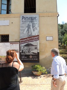 Sulla facciata del Museo del Merletto, il manifesto della mostra fotograficadedicata al fotoreporter Enrico Valentini   -   Di spalle, a destra, il signore Italo Marri.